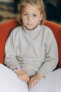 A young girl sitting indoors, reading a Braille book with focus and concentration.